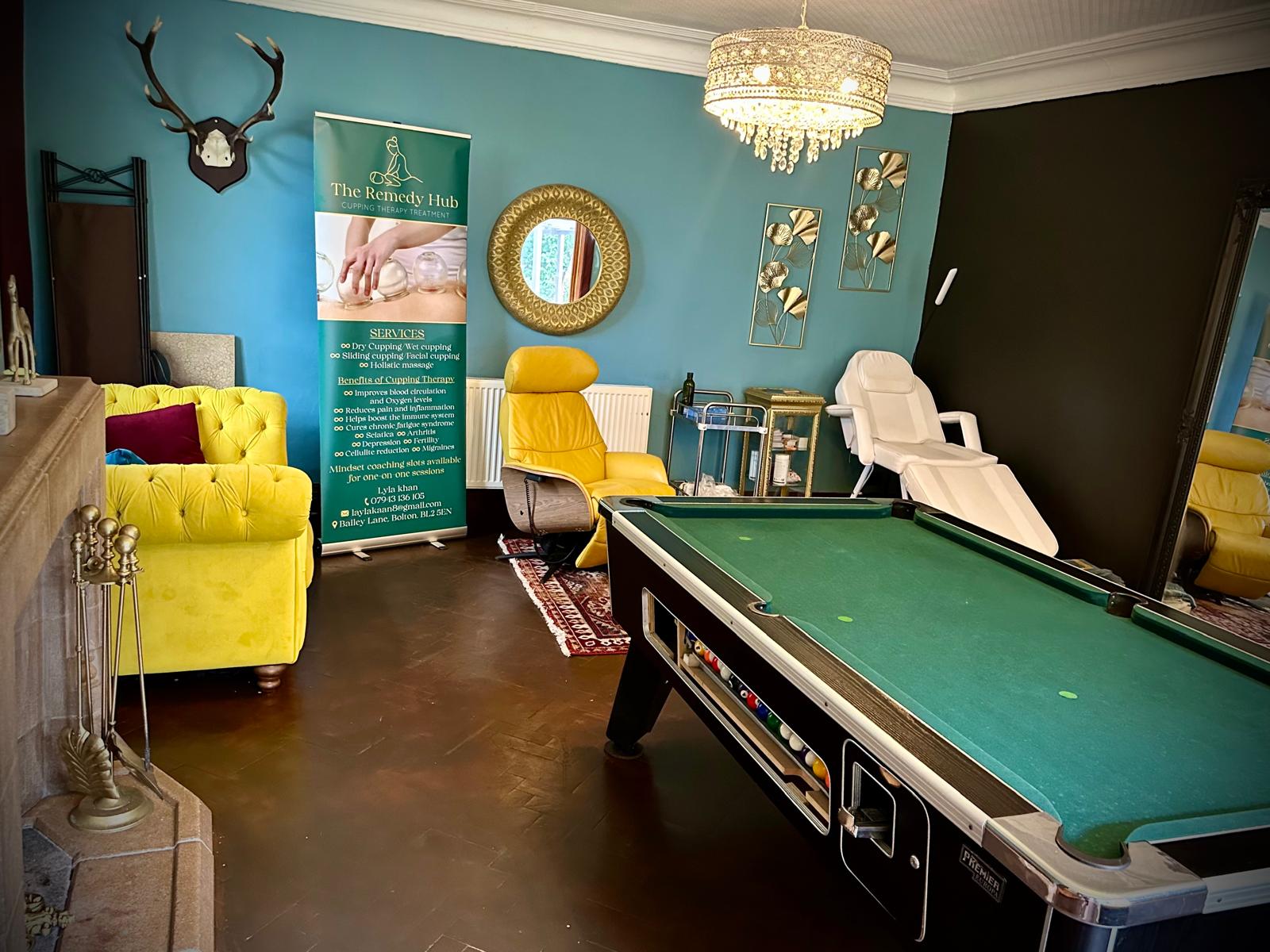A large indoor lounge or event space featuring a green pool table, a bright yellow tufted armchair, a white reclining chair, a blue wall, decorative gold wall art, and a hanging chandelier.